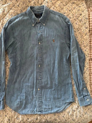 Jeansskjorta från Polo Ralph Lauren - Klassisk blå skjorta från Polo Ralph Lauren i slim fit strl M med button-down krage och broderad logga på bröstet. Skjortan har långa ärmar och knappar framtill. Tillverkad i mjuk bomull för en avslappnad men stilren look.