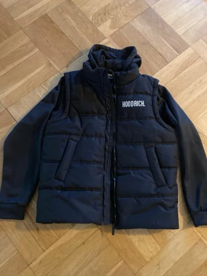 Svart västjacka från Hoodrich 13-15 år - Cool svart västjacka från Hoodrich med vit logotyp på bröstet och ryggen storlek 13-15 år. Jackan har en hög krage, luva, dragkedja framtill och två snedställda fickor. Materialet är quiltat och fylligt med tunnare material i ärmarna. Ärmarna är släta och jackan har en modern, sportig look. Nyskick