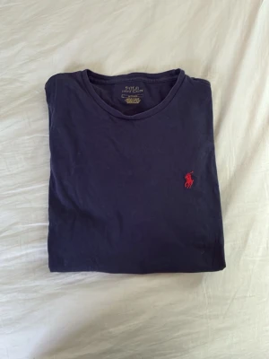 Marinblå t-shirt från Polo Ralph Lauren - Klassisk marinblå t-shirt från Polo Ralph Lauren med rund halsringning och den ikoniska röda logon broderad på bröstet. Tillverkad i mjuk bomull och har en enkel, stilren design som passar till allt.