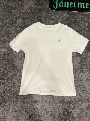 Vit t-shirt från Polo Ralph Lauren 14-16 år - Klassisk vit t-shirt från Polo Ralph Lauren med liten broderad logga på bröstet. T-shirten har rund halsringning och korta ärmar. Tillverkad i mjuk bomull som känns skön mot huden. Perfekt basplagg till garderoben.