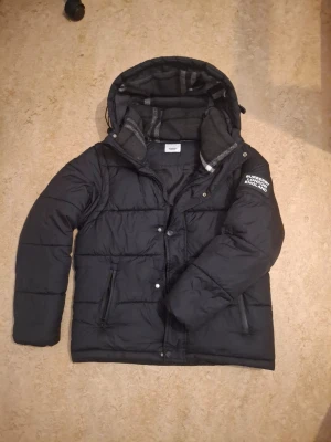 Svart pufferjacka från Burberry - Snygg svart pufferjacka som går och göra till en väst från Burberry med stor huva och mjukt rutigt innerfoder. Jackan har dragkedja, tryckknappar och två fickor framtill. På ärmen sitter en tydlig Burberry London England-logga i vitt. Perfekt för kalla vinterdagar. Det går även att glra till en väst för sommar dagar.
