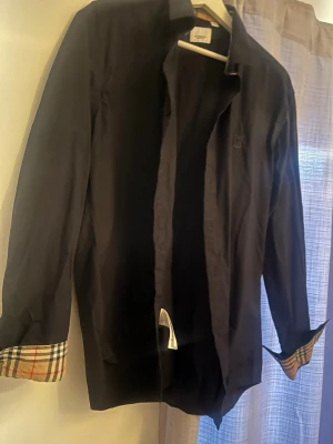 Svart skjorta från Burberry - Stilren svart skjorta från Burberry med klassiskt rutigt mönster på insidan av manschetterna. Skjortan har långa ärmar, knappar framtill och diskret Burberry-logga på bröstet. Passar även dig som har S 