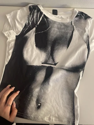 Vit t-shirt med grafiskt tryck - Unik vit t-shirt med svart grafiskt tryck som täcker hela framsidan. Trycket är i pop art-stil  och skapar en cool, edgy look. T-shirten har rund hals och korta ärmar. Perfekt för dig som vill sticka ut med din outfit.