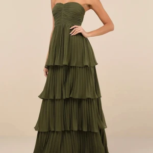Grön axelbandslös långklänning - Säljer en snygg axelbandslös långklänning i olivgrönt med flera lager volanger och plisserad kjol. Klänningen har en figurnära överdel och faller elegant ner till golvet. Perfekt för dig som vill sticka ut på nästa bal eller fest.