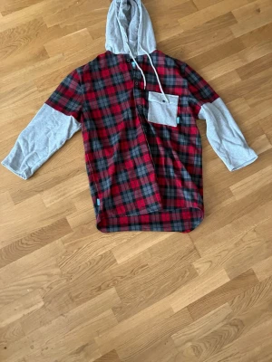 Rutig hoodie med grå ärmar och huva - Unik hoodie med röd och svart rutig kropp, grå ärmar och huva. Stor bröstficka i grått tyg med knapp, knäppning framtill och coolt rutigt mönster även på ryggen. Perfekt för dig som gillar streetwear och vill sticka ut. Mjuk bomullskänsla och avslappnad passform.