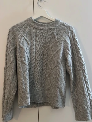 Grå kabelstickad tröja Gina Tricot - Säljer en grå kabelstickad tröja från Gina Tricot. Tröjan har rund halsringning, långa ärmar och ribbade muddar vid ärmslut och nederkant. Mönstret är klassiskt flätstickat och materialet känns mjukt och mysigt.