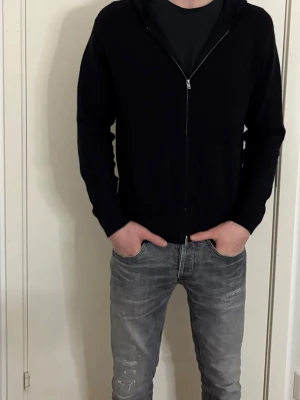 Svart hoodie med dragkedja - Säljer en svart hoodie med dragkedja och huva. Hoodien har en enkel och stilren design, perfekt för dig som gillar basic plagg. Materialet ser ut att vara mjukt och bekvämt, med långa ärmar och klassisk passform. Passar till jeans eller joggers.