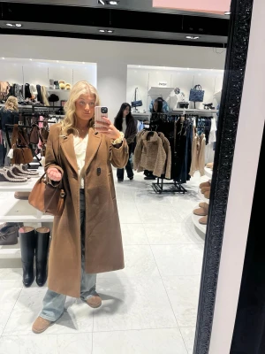 Kamelbrun lång kappa i ullmix - Stilren kamelbrun kappa med rak passform och klassisk krage. Kappan har knäppning framtill och är i ett mjukt ullblandat material. Perfekt för dig som gillar en tidlös och trendig look.