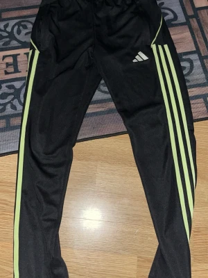 Svarta Adidas träningsbyxor - Säljer ett par svarta träningsbyxor från Adidas med de klassiska tre ljusgröna ränderna längs benen och Adidas-logga på ena låret. Byxorna har resår i midjan och är tillverkade i ett mjukt, stretchigt material som passar perfekt till sport och träning.