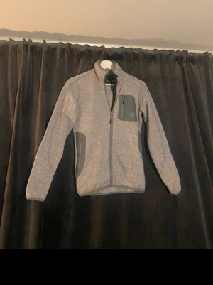 Ljusgrå fleecejacka  - Snygg ljusgrå fleecejacka med hel dragkedja framtill och praktiska fickor med grå detaljer. Jackan har hög krage och lång ärm, perfekt för lager på lager under kyliga dagar.