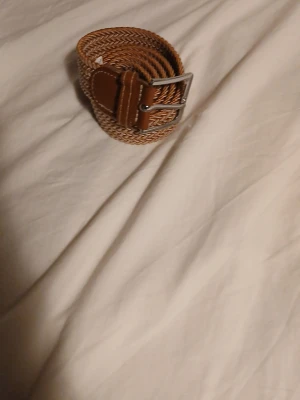 Flätat brunt bälte med silverfärgat spänne - Snyggt flätat bälte i brun färg med ett stilrent silverfärgat spänne. Bältet har en klassisk design och är gjort i ett material som liknar skinn. Perfekt för att lyfta vilken outfit som helst. Priset kan diskuteras.