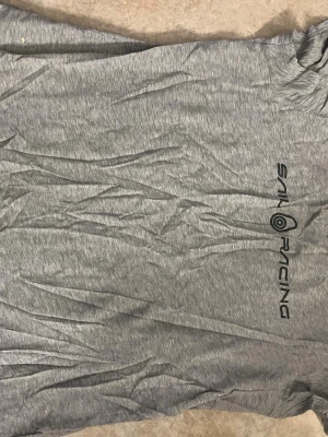 Grå t-shirt från Sail Racing - Snygg grå t-shirt från Sail Racing med tryckt logga på bröstet. Tillverkad i mjuk bomull och har en klassisk passform med korta ärmar. Perfekt till jeans eller shorts för en avslappnad look.