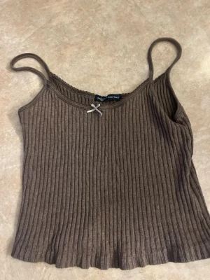 Brun ribbad linnetopp med rosett - Snygg brun ribbad linnetopp från Brandy Melville med tunna axelband och en liten söt rosett framtill. Perfekt till jeans eller kjol och superenkel att styla för en chill look.