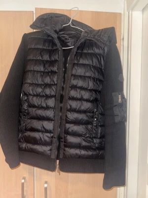 Svart pufferjacka från Moncler - Säljer en svart pufferjacka från Moncler med stickade ärmar och quiltad front. Jackan har hög krage, dragkedja framtill och fickor med tryckknappar. Detaljerad med en liten ficka på ärmen och klassisk Moncler-logga på insidan. Perfekt för kalla vinterdagar.har två små skador