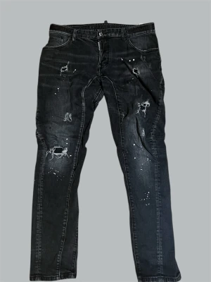 DSQUARED2 Svarta Jeans - DSQUARED2 svarta jeans med distressed-design, flera slitningar och ripped-detaljer på låren och knäna samt vita färgstänk som ger en rå streetlook. Klassisk femficksmodell i mörk tvätt. Litet slitage i Jeansknappen. 