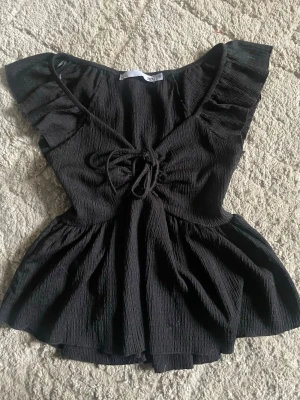 Svart volangtopp från 157 - Supersöt svart topp från 157 med volangdetaljer vid axlarna och knyt framtill. Toppen har en lätt rynkad struktur och v-ringad hals. Perfekt att styla med jeans eller kjol för en trendig look.