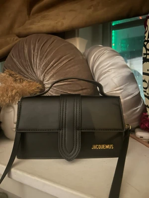 Svart  handväska i läder - Säljer en svart handväska  i  slätt läder med guldfärgad logga på framsidan. Väskan har ett stilrent, rektangulärt format lock och justerbar axelrem. Perfekt för dig som gillar minimalistisk och trendig design.