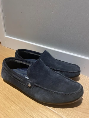 Mörkblå mocka loafers från Next - Snygga mörkblå loafers i mocka från Next med klassisk design och diskret detalj på sidan. Skorna har en mjuk ovandel och räfflad sula för extra grepp. Perfekta för en stilren och avslappnad look.