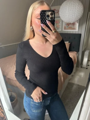 Svart ribbad långärmad topp - Svart långärmad topp med v-ringning och ribbad struktur. Sitter tight och har en klassisk, stilren look som passar till mycket. Perfekt att matcha med jeans eller kjol.