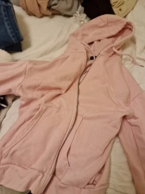 Ljusrosa hoodie med dragkedja 157 - Ljusrosa hoodie från 157 med dragkedja framtill och två stora fickor. Tröjan har huva med dragsko och ribbade muddar vid ärmslut och nederkant. Perfekt för en avslappnad och chill stil. Materialet känns mjukt och bekvämt, troligen bomullsblandning. Passar s och xs