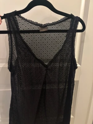 Svart spetslinne från vero moda - Säljer ett svart spets linne i märket GOSHA från vero Moda i transparent spetsmaterial med fint mönster. Linnet har v-ringning och spetskant runt halsen och ärmlösa axlar. Perfekt för att styla lager på lager eller under en kavaj.