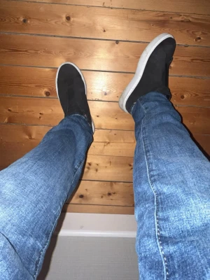 Svarta slip-on sneakers med vit sula - Säljer ett par svarta slip-on sneakers med tjock vit sula. Skorna har en minimalistisk design utan synliga snören och är tillverkade i ett tygmaterial. Perfekta för dig som gillar enkel och stilren look.