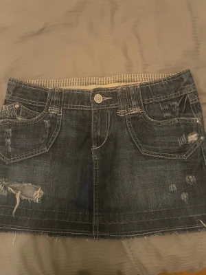 Mörk jeanskjol med slitningar - Säljer en mörkblå jeanskjol i kort modell med slitna detaljer och rå kant nertill. Kjolen har fem fickor, synliga sömmar och en knapp framtill. Perfekt för dig som gillar en edgy och avslappnad stil. 