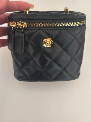 Svart quiltad handväska med guldkedja - Snygg svart quiltad handväska med guldfärgad kedja och dragkedja. Väskan har en liten guldfärgad blomdetalj framtill och är tillverkad i vadderat konstläder