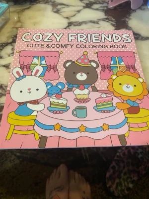 Cozy Friends målarbok - En söt och mysig målarbok med titeln Cozy Friends. Omslaget är rosa med gulliga djurfigurer som kanin, björn och lejon som fikar tillsammans. Boken har flera färgglada och svartvita illustrationer att färglägga, perfekt för dig som gillar kawaii och pyssel.