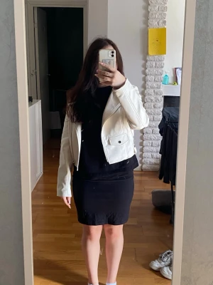 Vit skinnjacka i bikerstil - Fräsch vit skinnjacka i kort modell med klassisk bikerkrage och silvriga dragkedjor. Jackan har två fickor med lock framtill och en kortare passform. Perfekt för dig som vill ha en stilren och trendig look. Egentligen storlek M men passar bättre som XS. Har katter hemma!!!