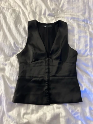 Svart satinväst från Zara - Stilren svart väst från Zara i glansigt satinmaterial. Västen har djup v-ringning och knappar framtill för en snygg och modern look.