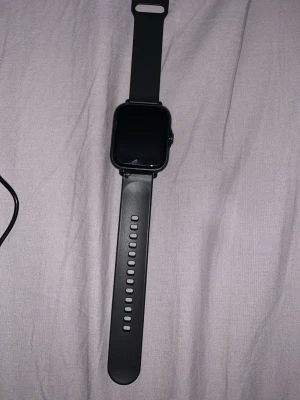 Svart smartklocka med silikonarmband - Stilren svart smartklocka med fyrkantig display och mjukt silikonarmband. Klockan har en rund röd knapp på sidan och justerbart spänne för perfekt passform. Passar dig som vill ha koll på notiser och hälsa direkt på handleden.