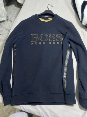 Svart Hugo Boss hoodie med logga. Den passar M o L - Snygg svart hoodie från Hugo Boss med stor guldfärgad BOSS-logga och texten HUGO BOSS på bröstet. Har dekorativa band med BOSS-tryck längs sidorna. Klassisk passform med rund hals och ribbade muddar. Perfekt för en trendig och avslappnad stil.