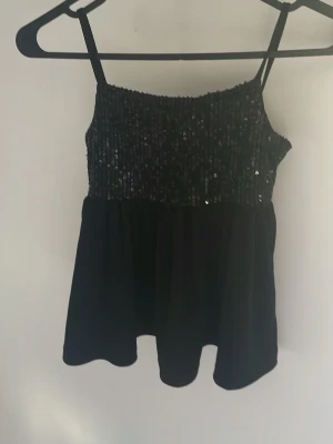 Svart glittrig linnetopp med paljetter - Säljer en svart linnetopp med smala axelband och glittrig paljettdekor upptill. Toppen har en luftig passform och är perfekt för dig som vill sticka ut med lite extra shine. Passar till många olika outfits och är riktigt snygg!