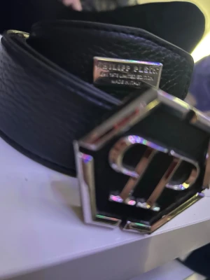 Svart skärp från Philipp Plein - Snyggt svart skärp från Philipp Plein med en stor, sexkantig metallspänne med qp-logga. Skärpet är gjort i präglat skinn och har en exklusiv känsla med metallplatta vid sidan. Perfekt för att lyfta vilken outfit som helst.