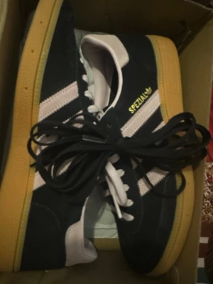 Adidas Spezial svarta sneakers - Säljer ett par Adidas Spezial sneakers i svart mocka med vita detaljer och klassiska tre ränder på sidorna. Skorna har gulbruna gummisulor och vita skosnören. Adidas-logga och Spezial-tryck i gult på sidan. Perfekta för dig som gillar retrostil och streetwear.
