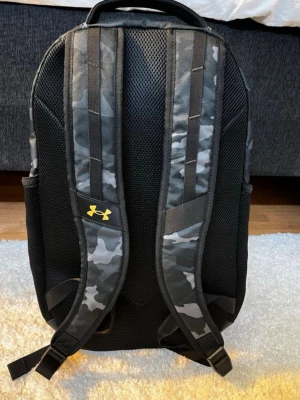 Under Armour camo ryggsäck - Ryggsäck från Under Armour i svart och grått camomönster med flera fack och vadderade axelremmar. Stor gul logga framtill och praktiskt bärhandtag upptill. Tillverkad i slitstarkt syntetmaterial.