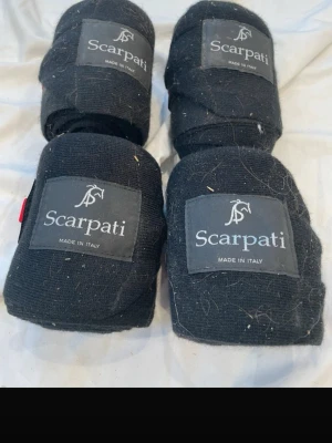 Svarta benlindor från Scarpati - Fyra svarta benlindor från Scarpati, tillverkade i Italien. De är gjorda i ett elastiskt tygmaterial och har kardborrestängning för enkel användning. Perfekta för hästträning eller transport. Klassisk design med Scarpati-logga påsydd på varje linda. Aldrig använda, men legat i ett skåp så de har blivit smutsiga och dammiga.