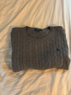 Grå kabelstickad tröja Polo Ralph Lauren - Klassisk grå kabelstickad tröja från Polo Ralph Lauren med rund halsringning och diskret broderad logga på bröstet. Tröjan har långa ärmar och ribbade muddar vid ärmslut och nederkant. Perfekt för lager på lager och stilren vardagslook.