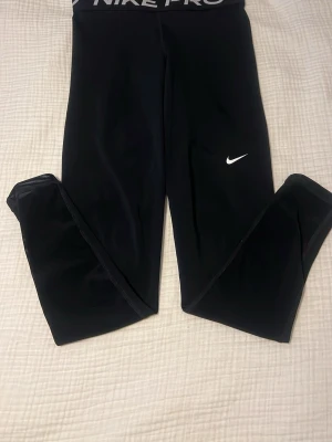 Svarta Nike Pro leggings - Säljer ett par svarta Nike Pro leggings med hög midja och elastisk linning där det står 'NIKE PRO'. De har en tight passform och är tillverkade i ett stretchigt funktionsmaterial. Klassisk vit Nike-logga på benet. Perfekta för träning eller chill.