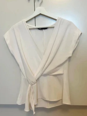 Vit omlottblus med knytband - Stilren vit omlottblus med djup V-ringning och knytband i midjan. Blusen har korta ärmar och är tillverkad i ett mjukt, lätt tyg som ger ett snyggt fall. Perfekt för en trendig och avslappnad look.