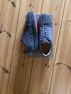 Gråa Louboutin sneakers med nitar - Säljer ett par gråa sneakers från Christian Louboutin i mocka med svarta nitar på tån och klassisk röd sula. Skorna har snörning och låg profil. Perfekta för dig som vill sticka ut med en edgy och lyxig vibe.
