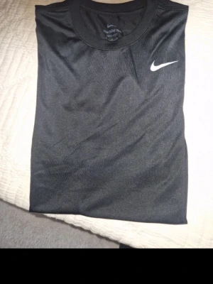 Svart Dri-Fit t-shirt från Nike - Snygg svart t-shirt från Nike med klassisk vit swoosh på bröstet. Tillverkad i Dri-Fit material som andas och håller dig torr. Perfekt för träning eller chill dagar. Enkel och stilren design med rund hals och korta ärmar.