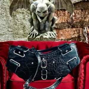 Svart väska i alternativ stil! - Svart gothic/alternative handväska i krokodilmönster och rockiga detaljer. Både handtag, avtagbart & justerbart axelband samt ett stort fack med dragkedja. B: 30 cm H: 15 cm. D: 10 cm. Oanvänd.