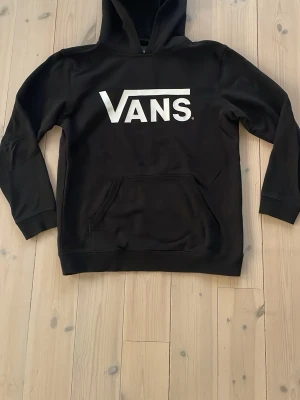 Svart Vans hoodie - Svart hoodie från Vans i storlek XL i junior storlek, passar även till storlek cirka 160-170 men kanske också 150 beroende på hur man vill ha det. Stor vit Vans-logga på bröstet. Klassisk modell med huva och känguruficka framtill. Tillverkad i mjukt material som passar perfekt för en avslappnad streetstil. Super fint skick och bra kvalitet.