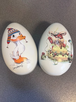 Påskägg i plåt av Lasse Åberg - Två vita påskägg i plåt med motiv av Lasse Åberg. Ena ägget har en anka med mössa och brev, det andra en mus med tårta och presenter. Perfekta som dekoration eller godisgömma till påsken. Samlarobjekt för dig som gillar Åbergs illustrationer.