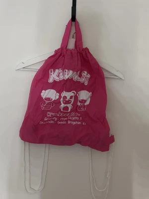 Rosa Kawaii ryggsäck med tryck - Säljer en rosa ryggsäck från Kawaii med vitt tryck av söta figurer och text. Väskan har vita snören som axelband och är tillverkad i lätt syntetmaterial. Perfekt för dig som gillar gulliga accessoarer och vill sticka ut lite extra.