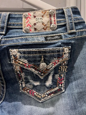 Mörkblå bootcut jeans från Miss Me - Snygga mörkblå bootcut jeans från Miss Me med broderade detaljer i rött, vitt och brunt på bakfickorna och vid linningen. Jeansen har slitningar och kontrastsömmar som ger en cool vintagekänsla. Perfekta för dig som gillar unik stil. Stor i storleken, känns som S om inte lite större! Ny pris 1000kr.