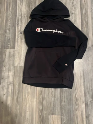 Svart hoodie från Champion - Svart hoodie från Champion med stor logga broderad på bröstet och en klassisk känguruficka framtill. Mjuk bomullsmix och huva med dragsko. Liten Champion-logga även på ärmen. Perfekt för en avslappnad och sportig stil. OPS den är solbränd!