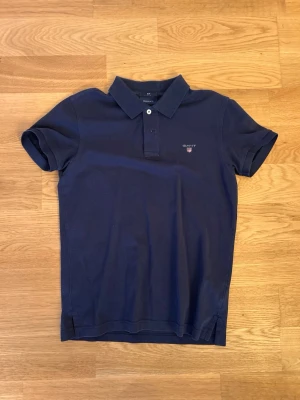 Pikétröja - Klassisk marinblå pikétröja från Gant med broderad logga på bröstet. Tröjan har korta ärmar, krage och knappar framtill. Slim fit-modell i mjuk bomull som är perfekt för en stilren look.